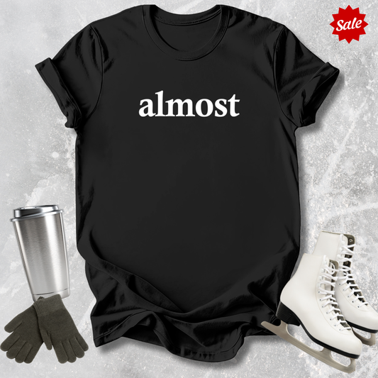 Black t-shirt with 'almost' text for figure skating coaches,, silver tumbler, black gloves, and white skates on a textured surface.