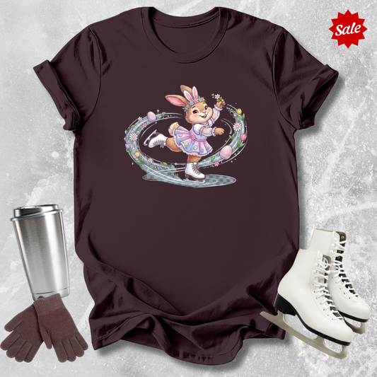 Brown t-shirt with a cartoon a figure skating Easter bunny design, next to a silver tumbler, brown gloves, and white ice skates on a textured gray background.