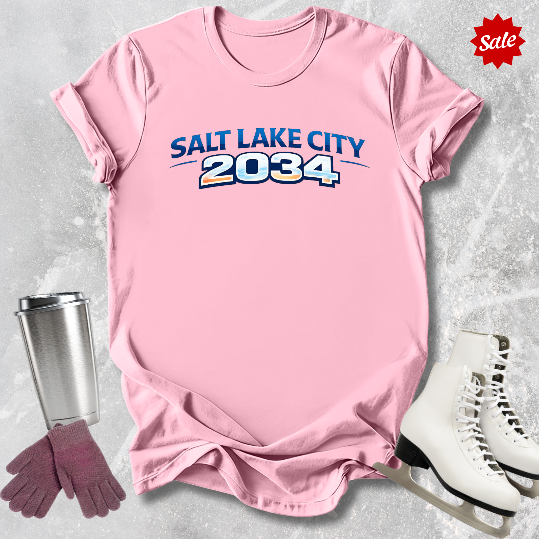 Pink t-shirt with 'Salt Lake City 2034' text, surrounded by a metal tumbler, gloves, and skates on a light gray background.