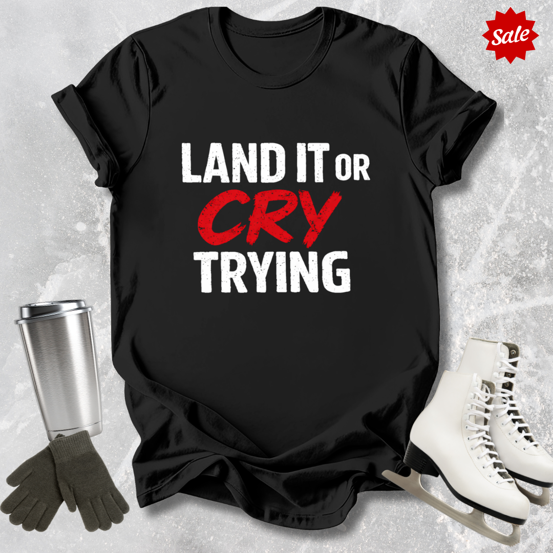 Black t-shirt with 'LAND IT OR CRY TRYING' text, next to a silver tumbler, black gloves, and white skates on a textured gray background.