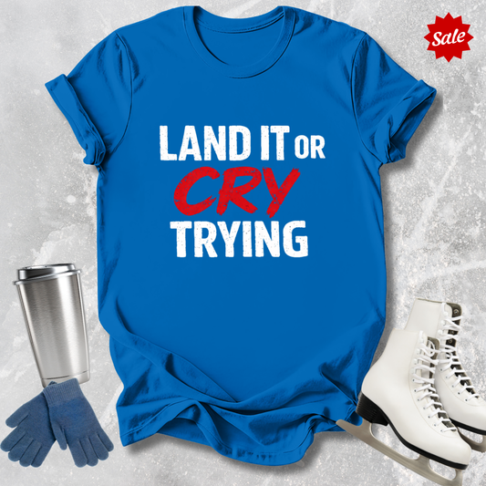 Blue t-shirt with 'LAND IT OR CRY TRYING' text, next to a silver tumbler, black gloves, and white skates on a textured gray background.
