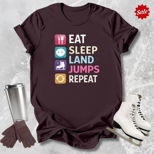 Brown t-shirt with 'Eat Sleep Land Jumps Repeat' text and graphics, next to a silver tumbler, gloves, and white skates on a textured gray background.