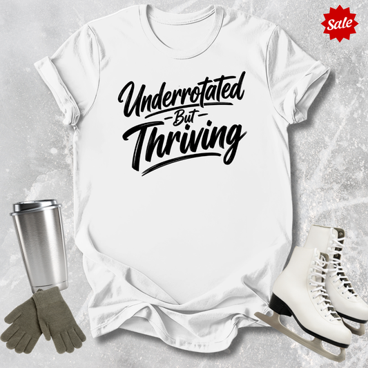 White t-shirt with 'Underrotated But Thriving' text, surrounded by a silver tumbler, gray gloves, and white skates on a textured surface.