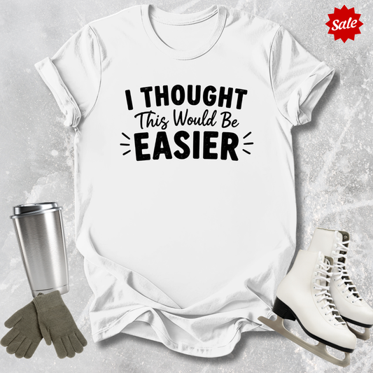 White t-shirt with 'I Thought This Would Be Easier' text, surrounded by ice skates, gloves, and a tumbler on a textured surface.