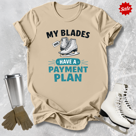Beige t-shirt with humorous ice skating graphic and text, surrounded by ice skates and gloves on a textured gray background.