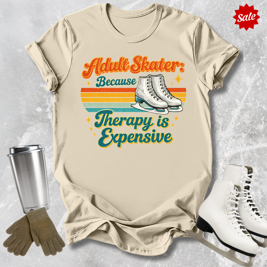 Beige t-shirt with 'Adult Skater: Because Therapy is Expensive' text, surrounded by ice skates, gloves, and a tumbler on a textured gray background.