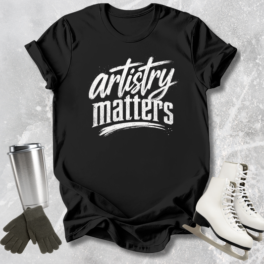 Black t-shirt with 'artistry matters' text, next to ice skates and a tumbler on a textured surface.