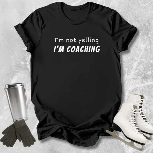 Black t-shirt with 'I'm not yelling I'm coaching' text, surrounded by ice skates and a tumbler on a textured surface.