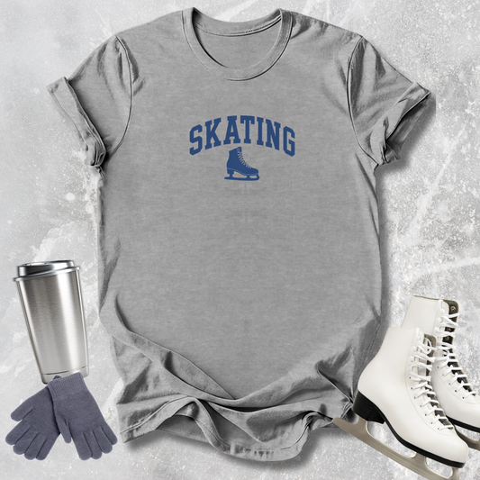 Gray t-shirt with 'SKATING' and ice skate graphic, next to a silver tumbler, gray gloves, and white skates on a textured surface.