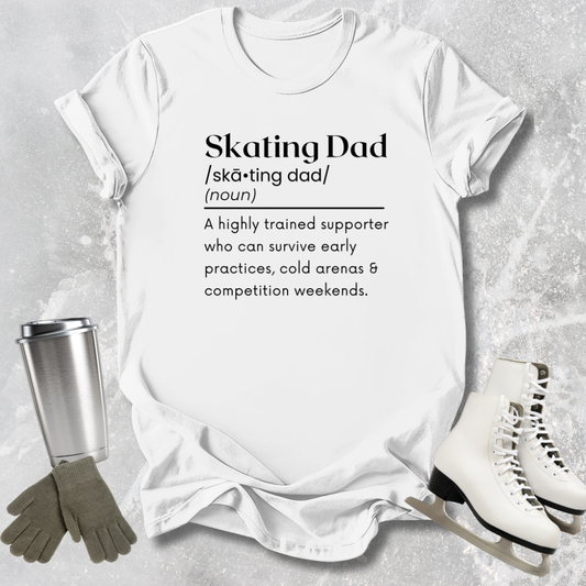 White t-shirt with 'Skating Dad' definition text, surrounded by ice skates, gloves, and a tumbler on a textured surface.