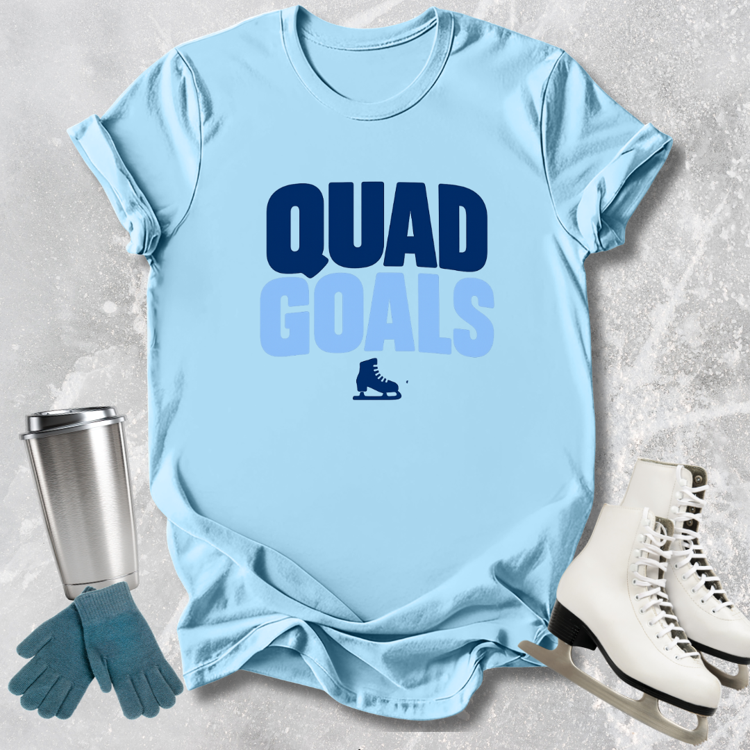 Light blue t-shirt with 'Quad Goals' text, ice skates, gloves, and skates on a textured surface