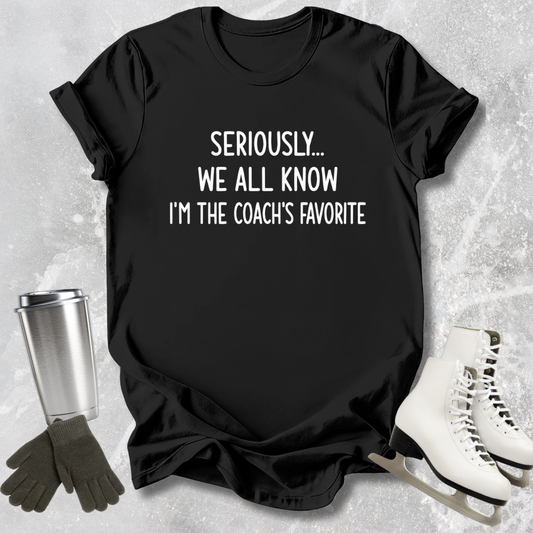 Black t-shirt with text 'Seriously... We all know I'm the coach's favorite' on a textured surface with accessories.