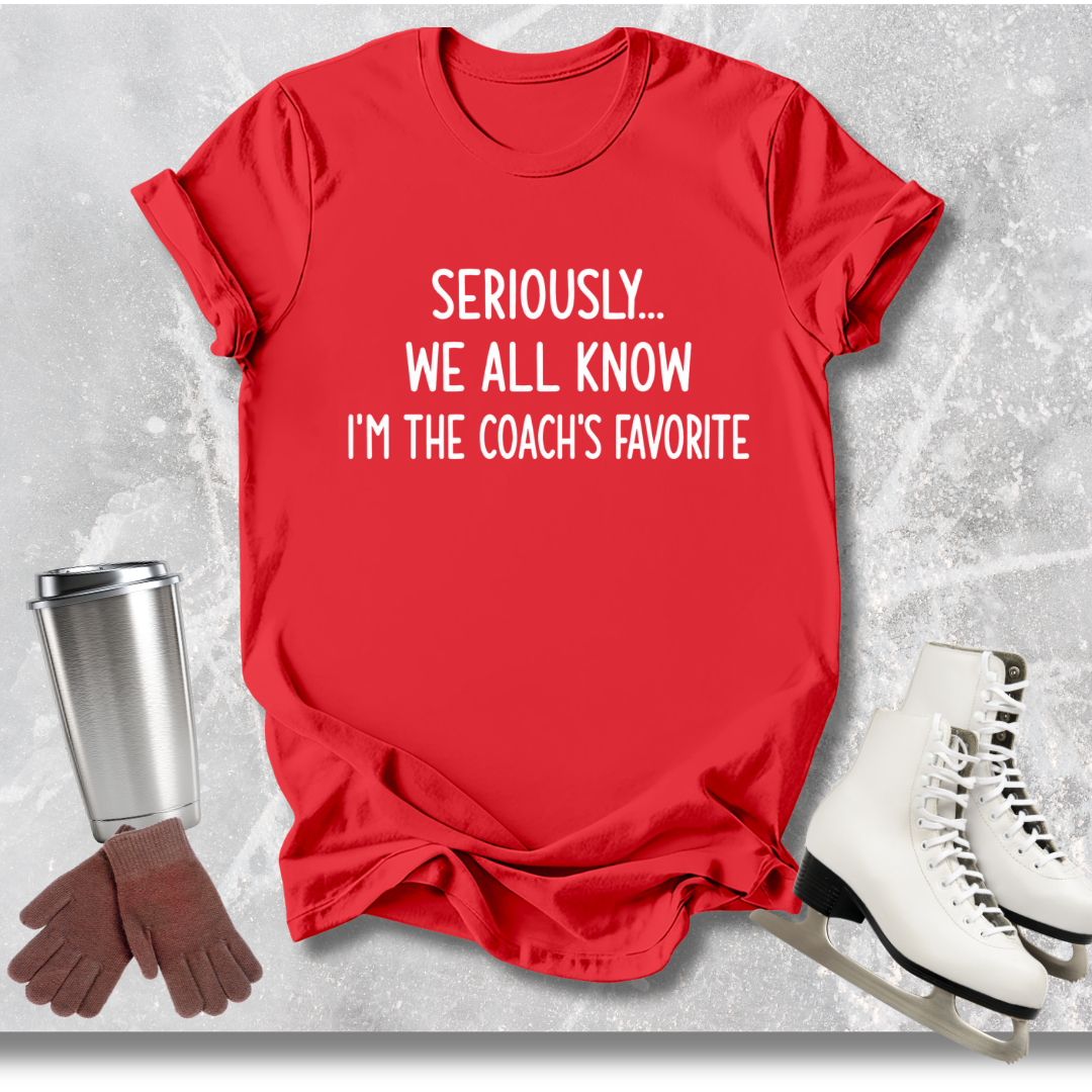 Red t-shirt with text 'Seriously... We all know I'm the coach's favorite' on a textured surface with accessories.