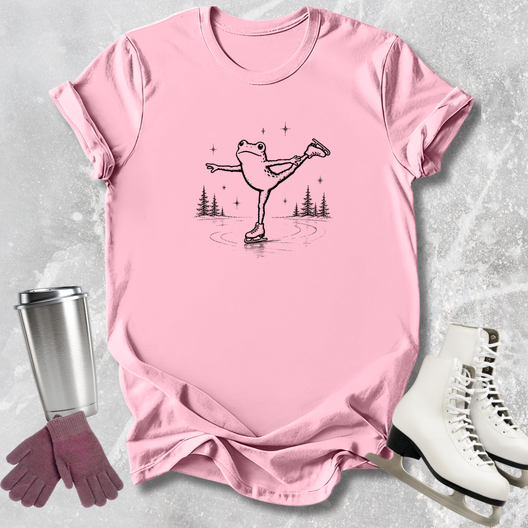 Pink t-shirt with a graphic of a frog ice skating, next to white ice skates and a silver tumbler on a light gray background.
