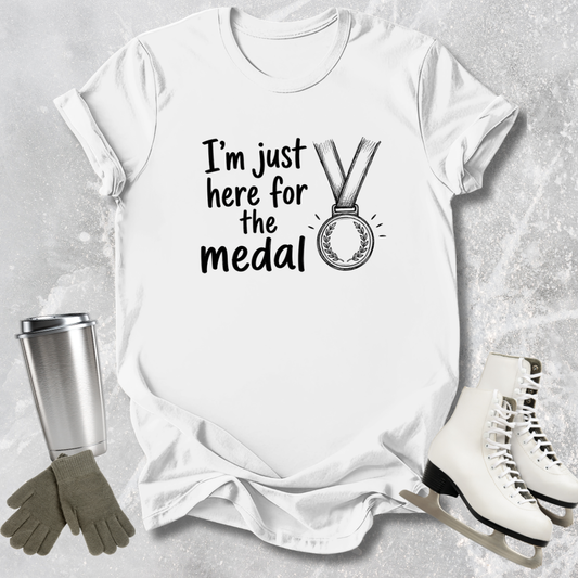 White t-shirt with 'I'm just here for the medal' text and graphic, surrounded by ice skates, gloves, and a tumbler on a light gray background.