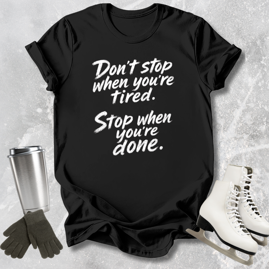 Black t-shirt with motivational quote, surrounded by a silver tumbler, black gloves, and white skates on a textured surface.