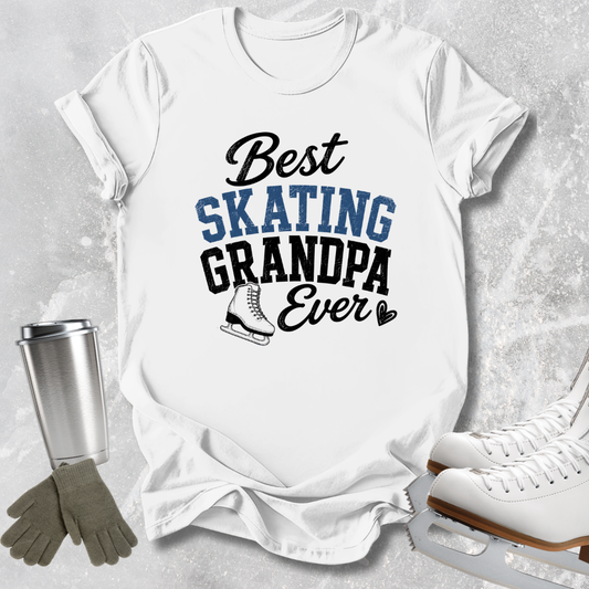 A white graphic T-shirt laid flat on a light gray textured background, with the text 'Best SKATING GRANDPA Ever' in black and blue lettering and a small ice skate illustration. A stainless steel tumbler, gray gloves, and white ice skates are arranged around the shirt as styled props.