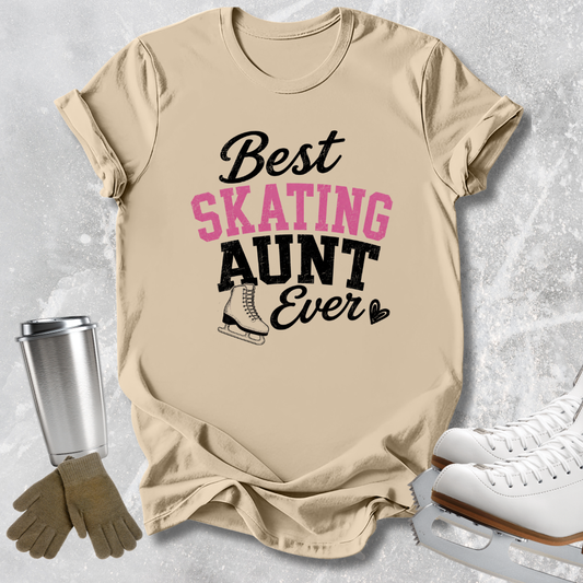 A beige graphic T-shirt laid flat on a light gray textured surface, with the text 'Best SKATING AUNT Ever' in black and pink lettering and a small ice skate illustration. White ice skates, tan gloves, and a metal travel tumbler are arranged around the shirt as styling props.