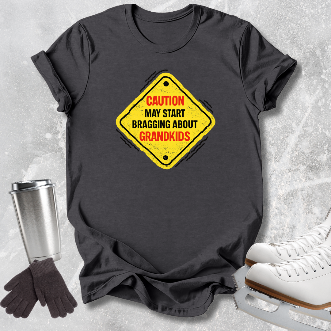 A dark charcoal gray t-shirt with a yellow diamond-shaped warning sign graphic that reads 'CAUTION MAY START BRAGGING ABOUT GRANDKIDS' in red and black text. The t-shirt is styled in a flat-lay arrangement on a light gray textured background, accompanied by a stainless steel travel mug, black gloves, and white ice skates.