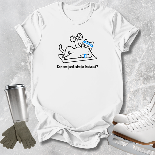 A white graphic t-shirt laid flat on a light gray textured surface, featuring a cartoon cat wearing a blue headband and lifting a dumbbell with the text 'Can we just skate instead?'. Ice skates, gloves, and a metal tumbler are styled around the shirt.