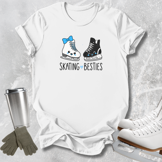 A white short-sleeve graphic T-shirt laid flat on a light gray textured surface, featuring two cartoon ice skates with faces and the text 'SKATING BESTIES.' A stainless steel tumbler, gray gloves, and white ice skates are placed around it as styling props.