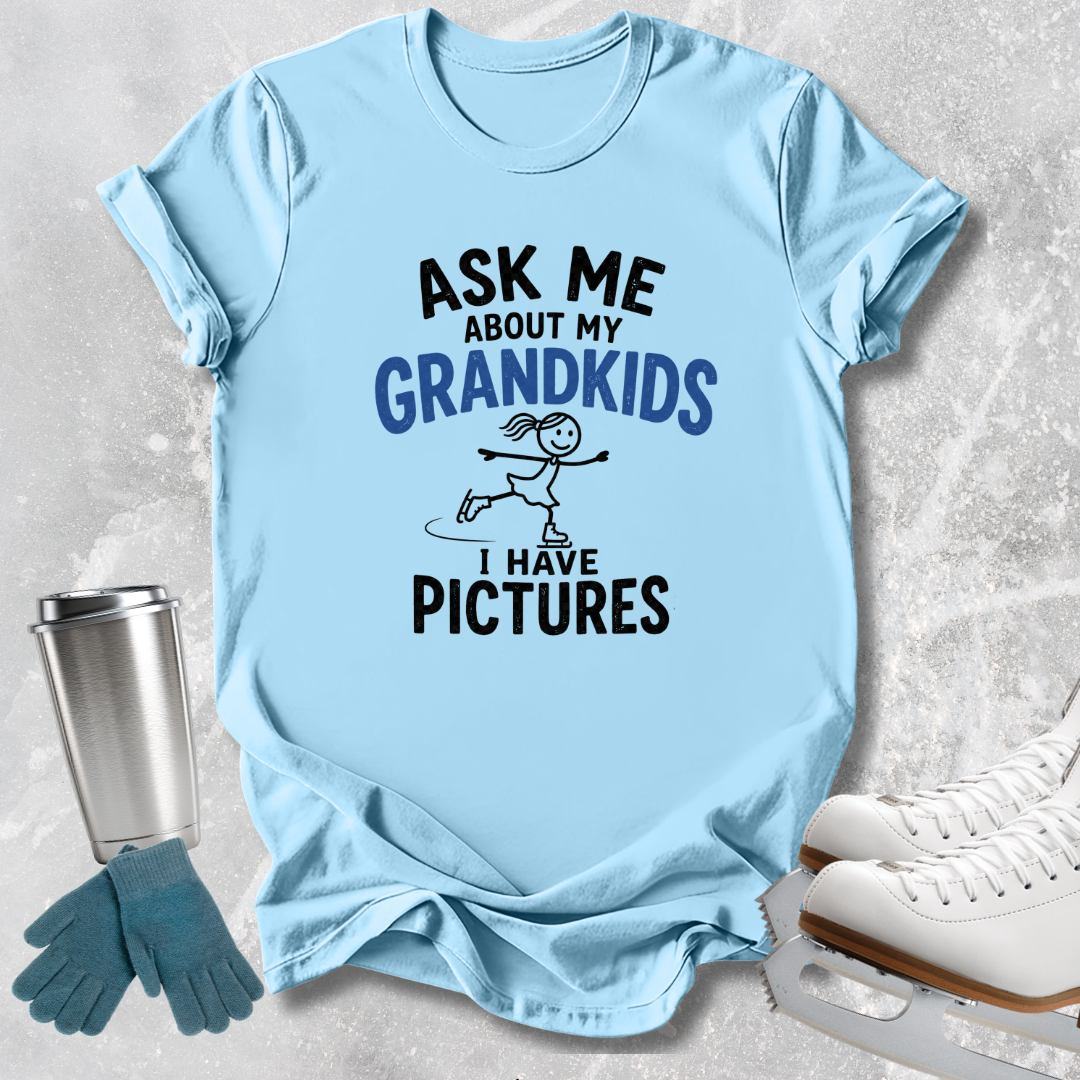 A light blue graphic t-shirt laid flat on a textured gray background, surrounded by ice skating props including white ice skates, blue gloves, and a silver tumbler. The t-shirt features the text 'ASK ME ABOUT MY GRANDKIDS I HAVE PICTURES' with a stick-figure skater graphic.