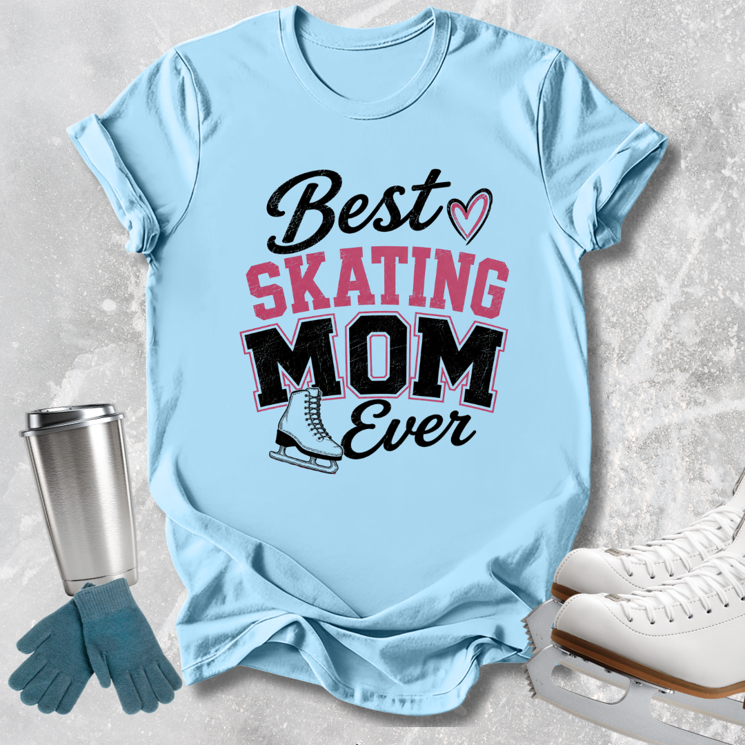 A light blue graphic T-shirt laid flat on a textured gray surface, with a printed design reading 'Best SKATING MOM Ever' in black and pink lettering and a small ice skate illustration. A stainless steel tumbler, blue gloves, and white ice skates are arranged around the shirt as props.