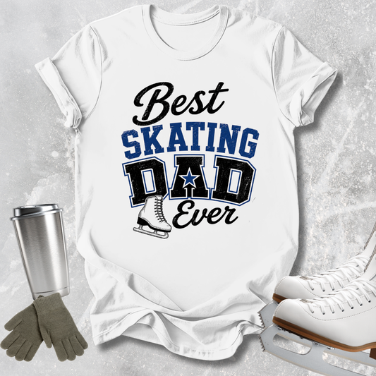 A white graphic t-shirt laid flat on a light gray textured surface, featuring the text 'Best SKATING DAD Ever' in black and blue lettering with a skater boot graphic. Ice skates, gloves, and a metal tumbler are arranged around the shirt as styling props.