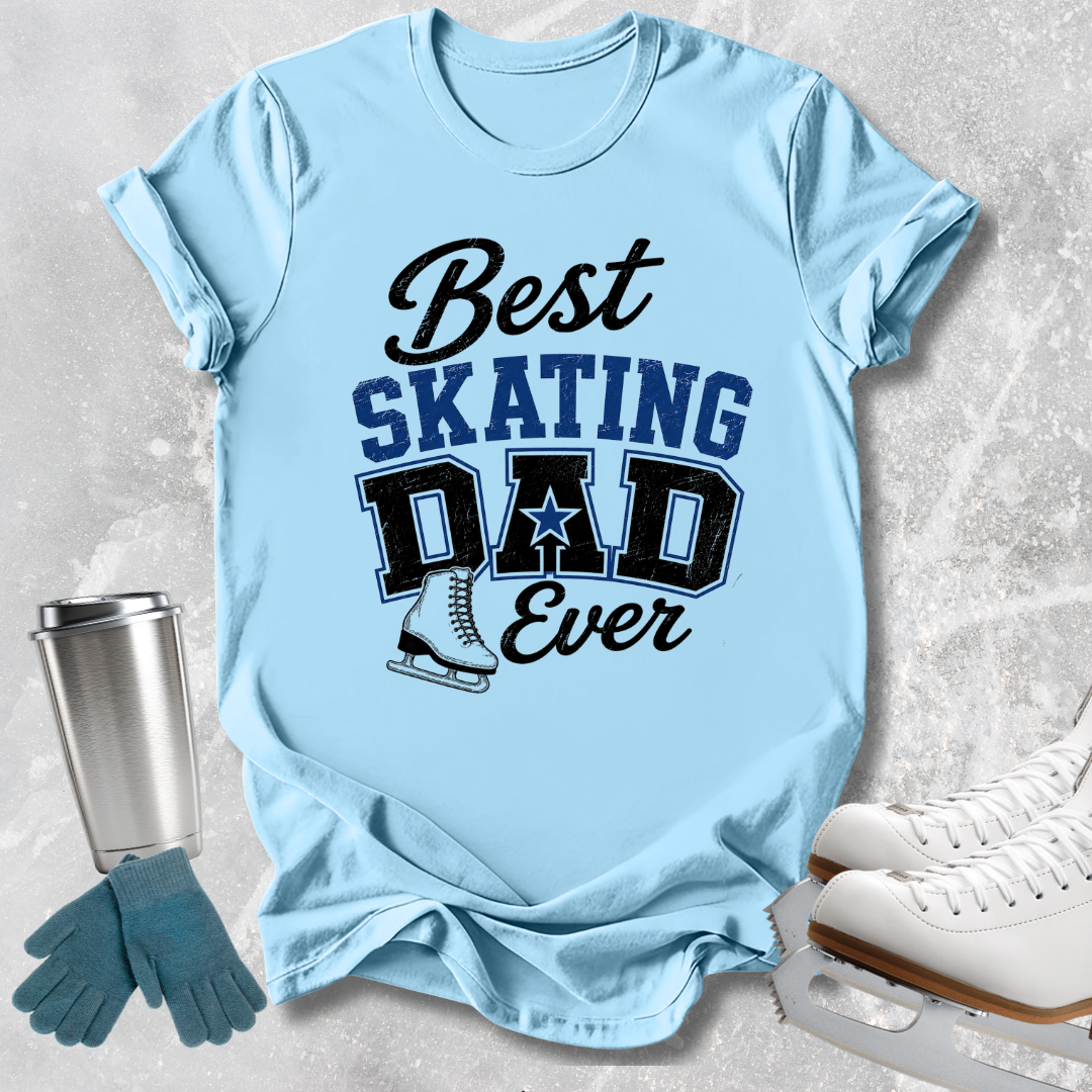 A light blue graphic T-shirt laid flat on a textured gray surface, with the words 'Best SKATING DAD Ever' in black and navy lettering and a small ice skate illustration. Ice skates, gloves, and a metal tumbler are styled around the shirt as props.