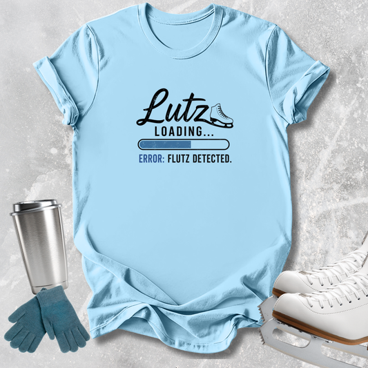A light blue graphic T-shirt laid flat on a light gray textured surface, with a stainless steel tumbler, teal gloves, and white ice skates around it. The shirt has a black printed design reading 'Lutz Loading.