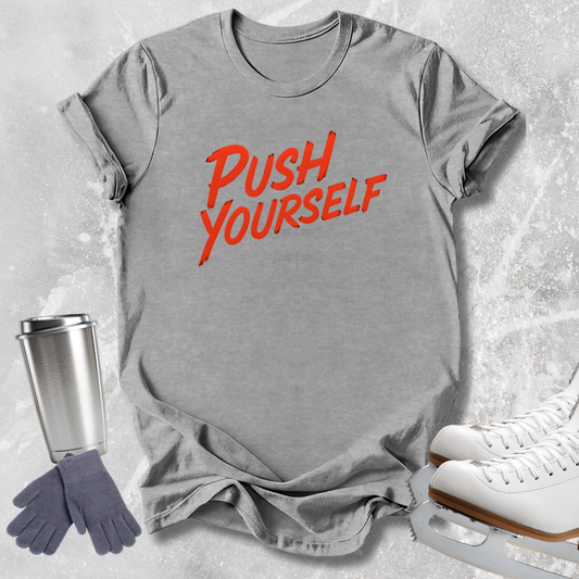 A heather gray short-sleeve T-shirt with the words 'PUSH YOURSELF' printed in red across the chest, styled in a flat lay with ice skates, gloves, and a travel tumbler on a light gray textured background.