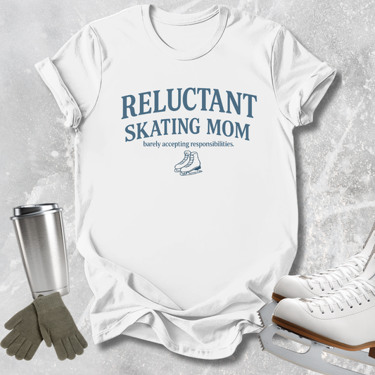 A white short-sleeve T-shirt laid flat on a light gray textured surface, printed with the phrase 'RELUCTANT SKATING MOM' and a small ice skate graphic in blue. Surrounding props include a stainless steel travel mug, gray gloves, and white ice skates.