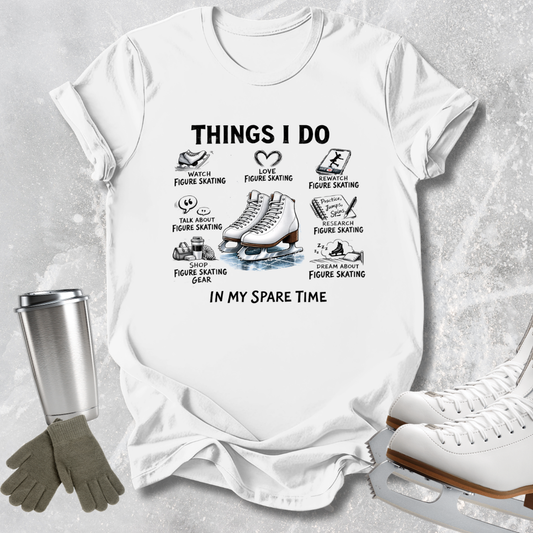A white graphic t-shirt laid flat on a light gray textured surface, featuring black text and illustrations related to figure skating. Surrounding props include a metal travel mug, gray gloves, and white ice skates.
