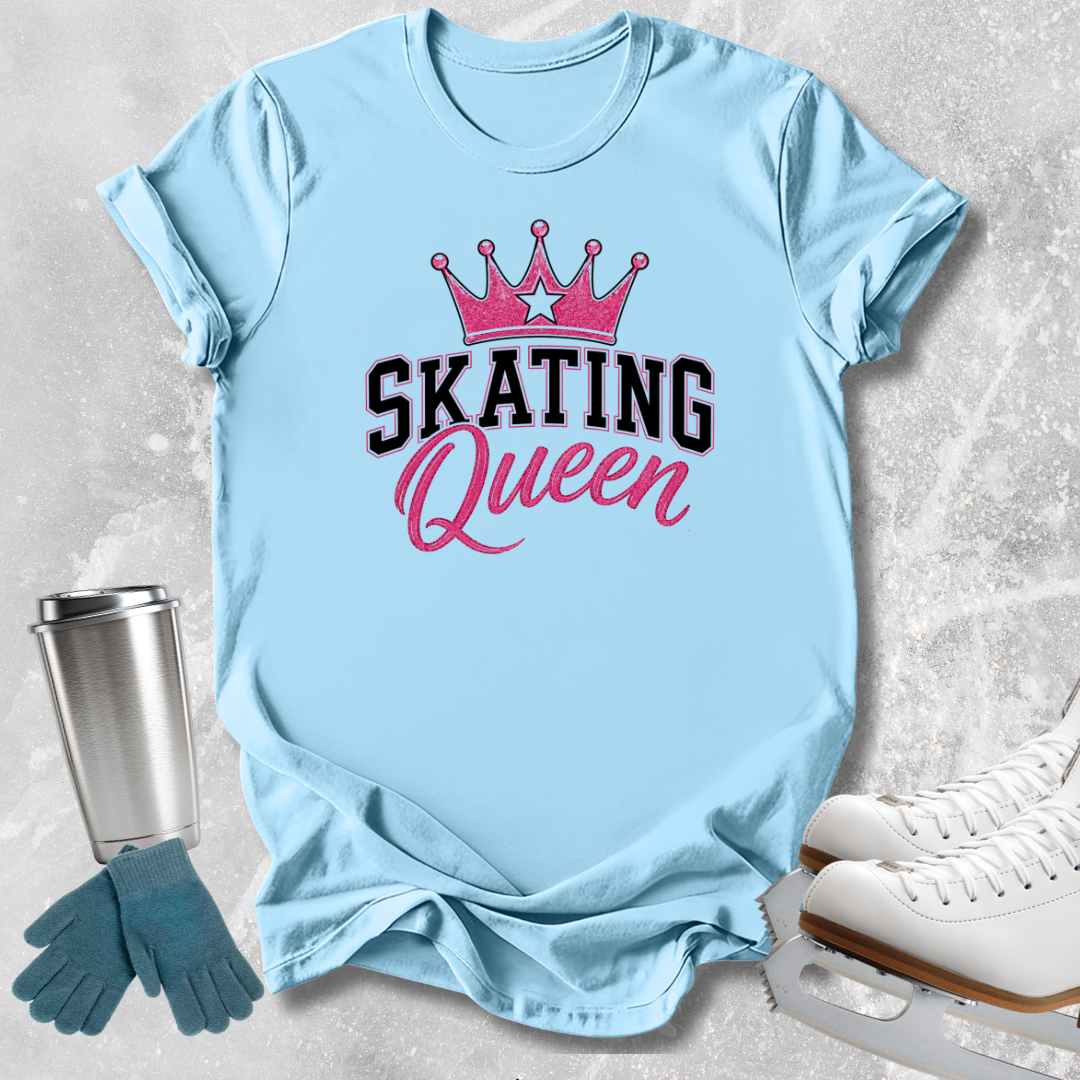 A light blue graphic T-shirt laid flat on a textured gray surface, featuring a pink crown above the words 'SKATING Queen' in black and pink lettering. Ice skates, gloves, and a metal tumbler are styled around the shirt as props.