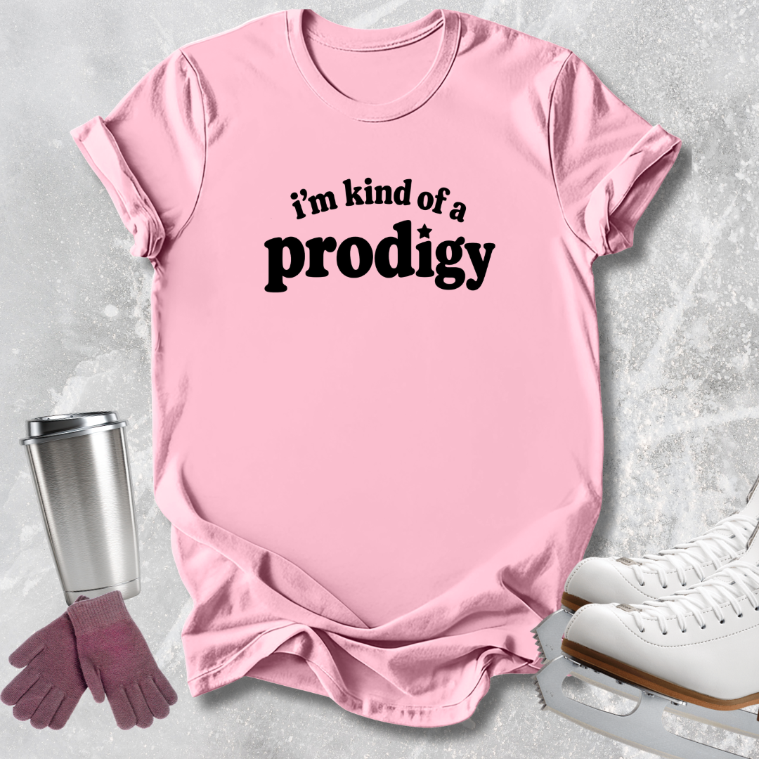 A light pink short-sleeve T-shirt with the phrase 'i'm kind of a prodigy' printed in black across the chest, styled in a flat lay with ice skates, gloves, and a travel tumbler on a light gray textured background.