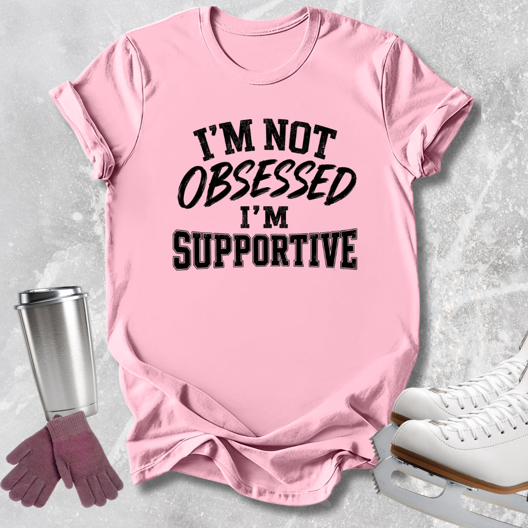 A pink short-sleeve graphic T-shirt laid flat on a light gray textured surface, with black text reading “I'M NOT OBSESSED I'M SUPPORTIVE.” Ice skate boots, gloves, and a metal cup are styled around the shirt as props.