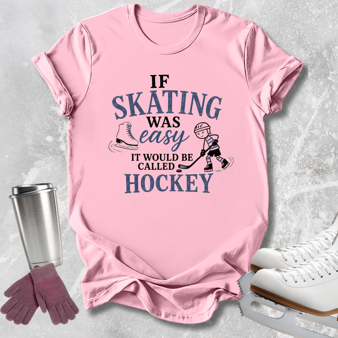 A pink short-sleeve graphic T-shirt laid flat on a light gray textured surface, with skating-themed props around it: a metal cup, gloves, and white ice skates. The shirt has a large printed slogan reading 'IF SKATING WAS EASY IT WOULD BE CALLED HOCKEY' in blue and black text with small skater and skate illustrations.