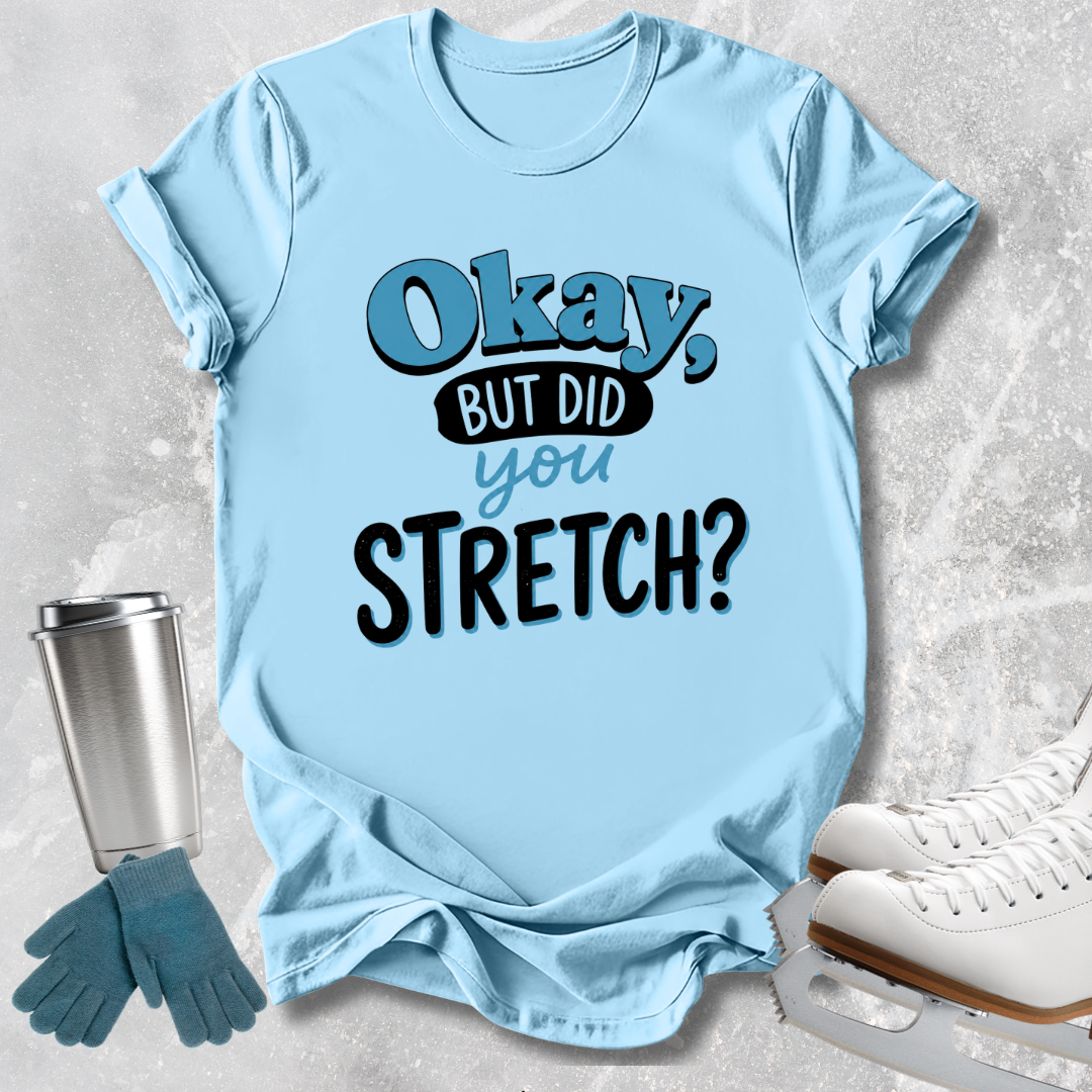 A light blue graphic t-shirt laid flat on a textured gray background, surrounded by ice skating props including white ice skates, blue gloves, and a stainless steel tumbler. The t-shirt features the text 'Okay, BUT DID you STRETCH?' in blue and black lettering.