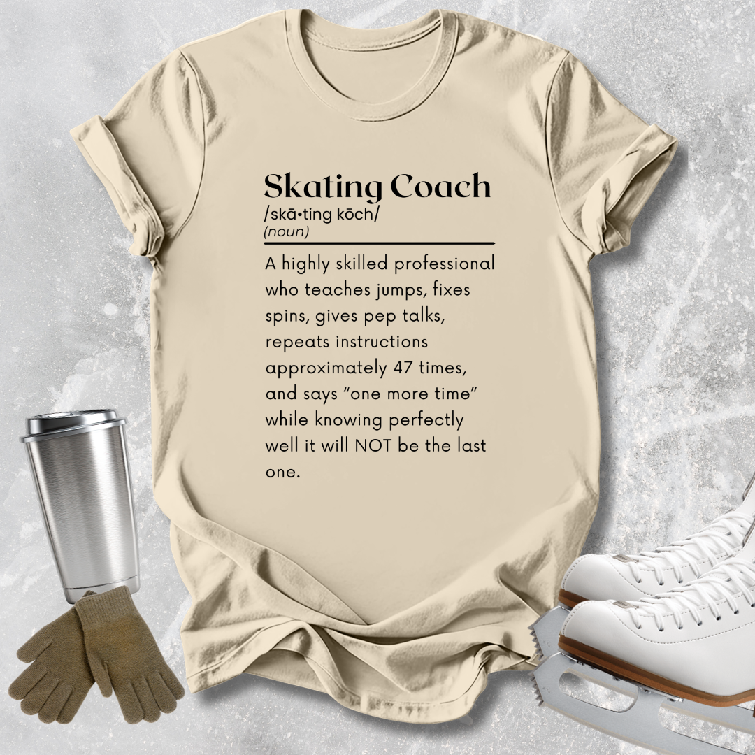 A beige graphic t-shirt featuring a humorous dictionary definition of a 'Skating Coach' printed in black text. The shirt is styled in a flat-lay arrangement on a textured gray background, accompanied by brown gloves, a silver travel mug, and white ice skates.