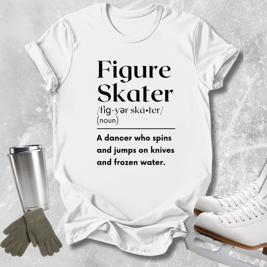 A white graphic T-shirt laid flat on a light gray textured surface, printed with the words 'Figure Skater' and a dictionary-style definition. Ice skates, gloves, and a metal cup are arranged around the shirt as styling props.
