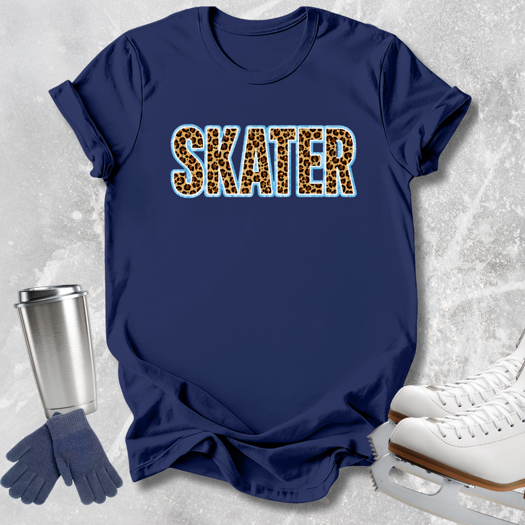 A navy blue short-sleeve T-shirt laid flat on a light gray textured background, with the word "SKATER" printed across the chest in leopard-print letters outlined in light blue. A silver travel mug, blue gloves, and white ice skates are arranged around the shirt as styled props.