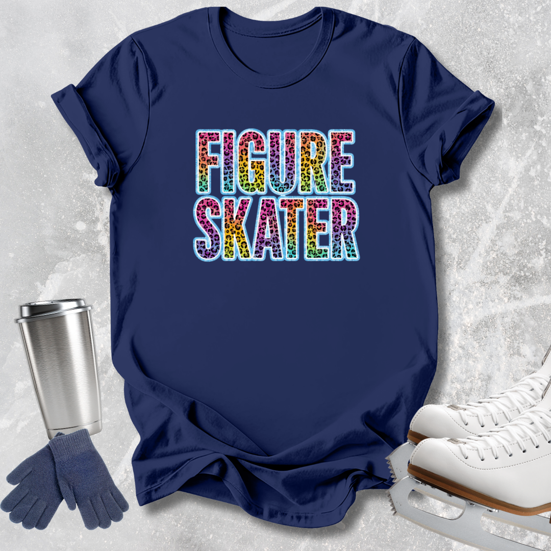 A navy blue graphic t-shirt with 'FIGURE SKATER' printed in colorful rainbow glitter-style letters, styled in a flat lay with ice skates, gloves, and a travel tumbler on a light gray textured background.