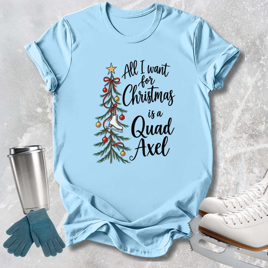 A light blue graphic T-shirt laid flat on a textured gray surface, with a Christmas tree illustration and the phrase 'All I want for Christmas is a Quad Axel' printed on the front. A stainless steel tumbler, blue gloves, and white ice skates are placed around it as styling props.