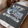 A dark gray blanket with a humorous graphic print is spread across a bed in a bedroom setting. The print features the text 'LIFE isn't all about SKATING & DOGS BUT IT SHOULD BE.