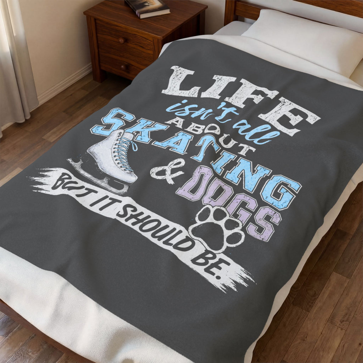 A dark gray blanket with a humorous graphic print is spread across a bed in a bedroom setting. The print features the text 'LIFE isn't all about SKATING & DOGS BUT IT SHOULD BE.