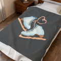 A dark gray blanket featuring a graphic of two light blue ice skates with a pink heart-shaped bow, shown on a bed in a bedroom setting.