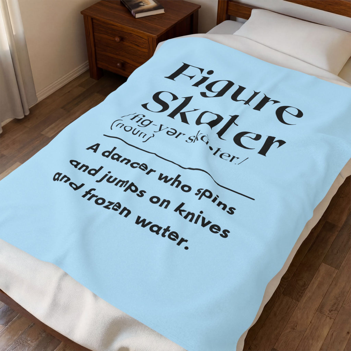 A light blue blanket with black printed text defining 'Figure skater' is draped over a bed in a bedroom setting.