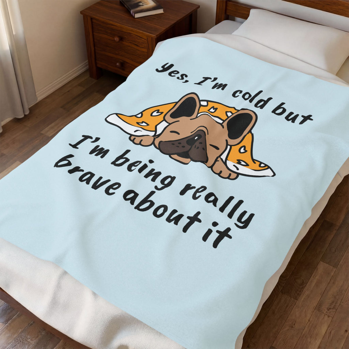 A light blue blanket with a cartoon French bulldog and the text 'Yes, I'm cold but I'm being really brave about it' shown on a bed in a bedroom setting.