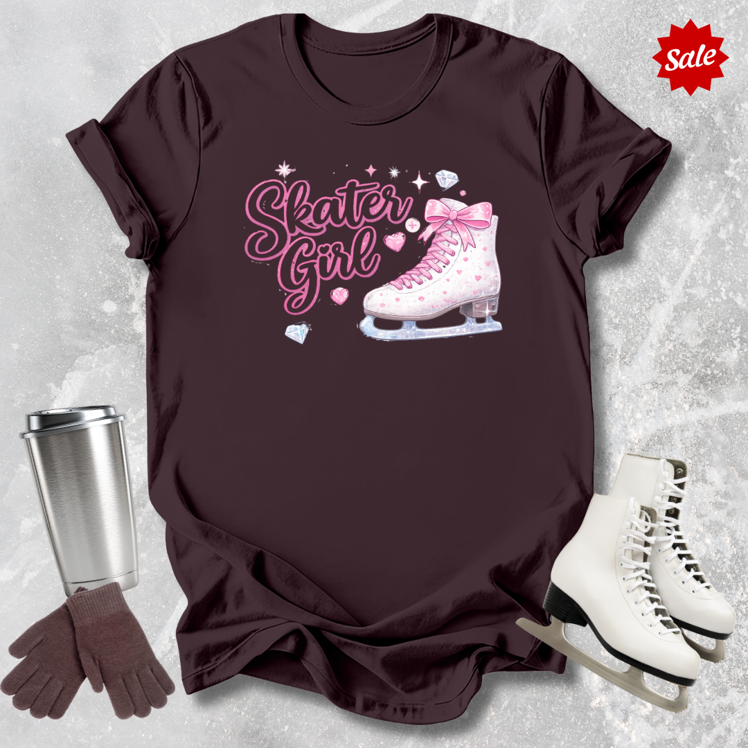 Brown t-shirt with 'Skater Girl' graphic, silver tumbler, brown gloves, and white ice skates on a textured gray background.
