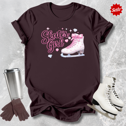 Brown t-shirt with 'Skater Girl' graphic, silver tumbler, brown gloves, and white ice skates on a textured gray background.
